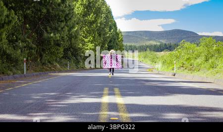 Una donna drappeggiata in un colorato poncho cammina lungo una strada di campagna deserta costeggiata da lussureggianti alberi verdi, circondata da pittoresche colline e un luminoso blu sk Foto Stock