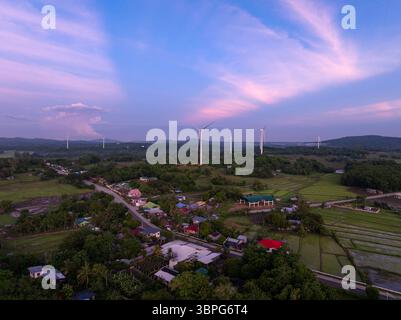 Vista aerea delle turbine eoliche alte in mezzo a una vegetazione lussureggiante, che gettano lunghe ombre mentre il sole tramonta a San Lorenzo, Guimaras, Filippine. Foto Stock