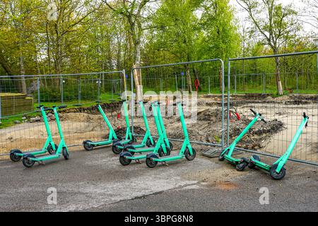 Scooter elettrici condivisi parcheggiati in un cantiere con recinzione di sicurezza e sfondo ad albero Foto Stock