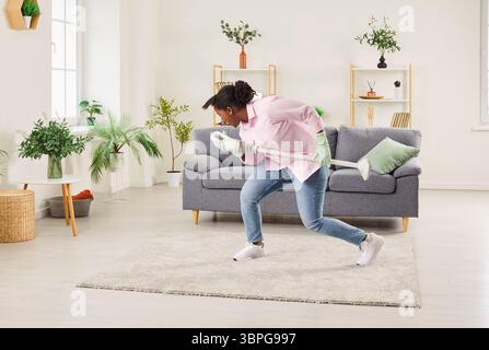 Donna afroamericana che usa l'aspirapolvere come microfono, divertendosi, ballando e cantando a casa. Foto Stock