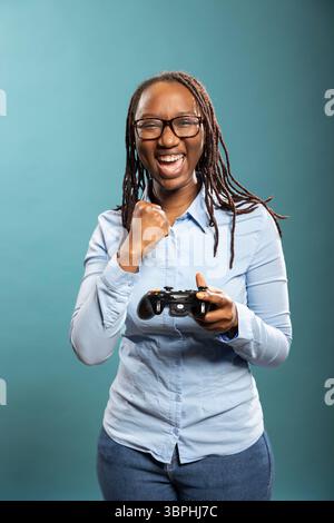 Donna afroamericana sorridente che tiene in mano il gamepad, divertiti con un divertente videogioco in uno studio con sfondo blu. Felice videogamer nero con controller moderno, che celebra una vittoria con pugno alzato. Foto Stock