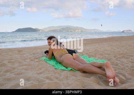 Donna felice in bikini che si rilassa e prende il sole sulla spiaggia sabbiosa con vista sull'oceano. Perfetto per vacanze estive e viaggi. Foto Stock