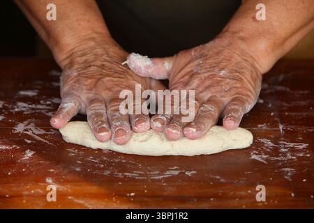 primo piano di mani di anziana donna, vestita in modo informale a casa, che fa impasto a mano su un semplice tavolo rustico di legno Foto Stock