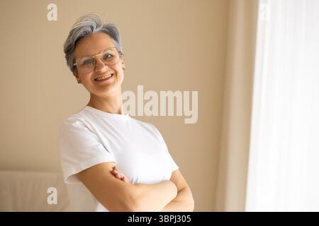 Una donna anziana, bella, anziana pensionata con i capelli grigi si trova vicino alla finestra, con le braccia incrociate. Foto Stock