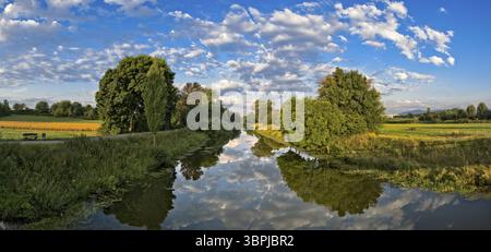 Letto del fiume Nidda in un'area ricreativa locale di Francoforte sul meno con diga, nuvole, cielo blu, alberi decidui sulla riva e campi che fiancheggiano Foto Stock