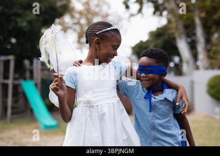 Fratelli afroamericani che indossano costumi da angelo e superman sorridenti e in piedi nel parco Foto Stock