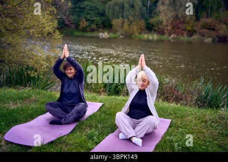 una donna di mezza età insegna alla sua vecchia madre esercizi somatici e yoga per mantenere la salute sul fiume in autunno Foto Stock