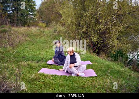 una donna di mezza età insegna alla sua vecchia madre esercizi somatici e yoga per mantenere la salute sul fiume in autunno Foto Stock