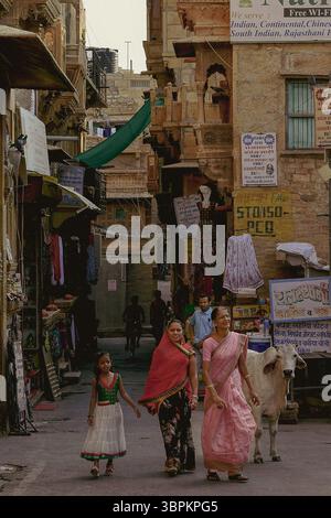 Tre donne indiane e una ragazza in abito tradizionale che camminano in una stradina stretta con negozi e una mucca, Jaisalmer, Rajasthan, India Foto Stock