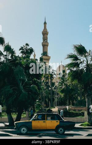 Taxi giallo e nero che passano davanti a una moschea circondata da palme ad Alessandria, in Egitto Foto Stock