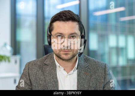 Ritratto ravvicinato di un giovane impiegato e assistente di ufficio di sesso maschile seduto sul posto di lavoro con una cuffia e guardando con attenzione la fotocamera. Foto Stock
