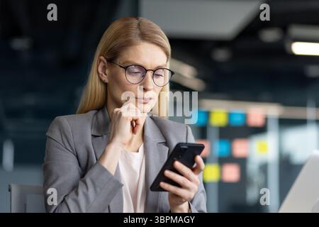 Una donna d'affari premurosa con un abito grigio è concentrata sul suo smartphone, con una mano sul mento. Foto Stock