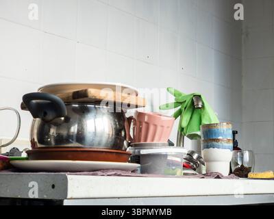 Un ripiano della cucina mostra una varietà di piatti sporchi impilati a caso insieme a prodotti per la pulizia e utensili. La luce naturale migliora la S. Foto Stock