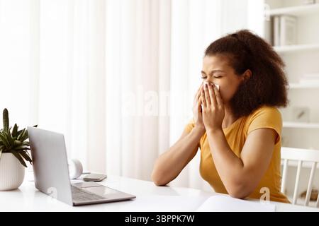 Giovane donna nera malata che starnutisce o soffia il naso mentre lavora da casa Foto Stock