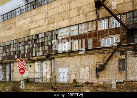 Edificio industriale abbandonato con finestre arrugginite, muri in mattoni e un cartello con la fermata del passaggio ferroviario Foto Stock