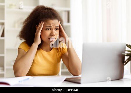 Giovane donna nera preoccupata che tiene la testa sotto stress mentre fissa il notebook Foto Stock