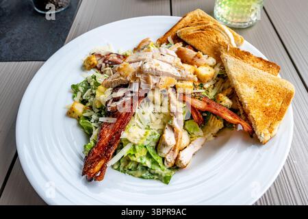 Porzione di insalata verde Caesar fresca con maionese, formaggio grattugiato e pezzetti di pollo, pane tostato arrosto in piatto bianco, cetriolo, primo piano. Foto Stock