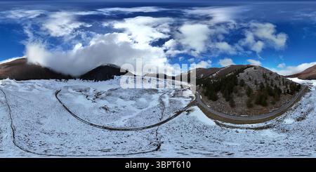 Un paesaggio invernale panoramico caratterizzato da colline innevate, una strada tortuosa e un cielo azzurro limpido con nuvole sparse. La scena cattura il sereno b Foto Stock
