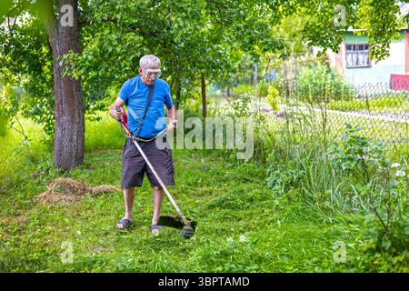 Uomo anziano che taglia l'erba coltivata con un decespugliatore. L'uomo anziano che indossa gli occhiali di protezione utilizza un tagliaerba per tagliare le erbacce sovracresciute in un giardino verde Foto Stock