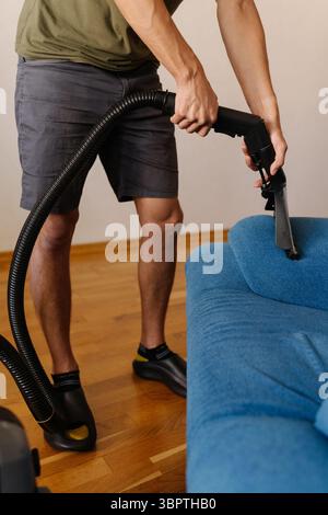 Colpo ritagliato verticale di giovane uomo che utilizza un moderno aspirapolvere per rimuovere sporco e polvere dal divano blu, garantendo una casa pulita e sana Foto Stock