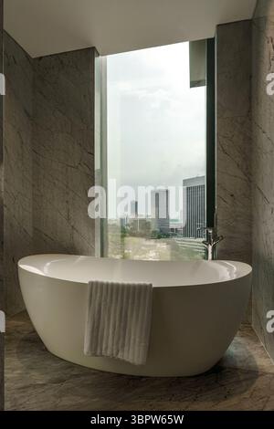 Vasca da bagno bianca indipendente minimalista in un lussuoso bagno in marmo grigio, posizionato da un'alta finestra con una splendida vista sullo skyline della città. Foto Stock
