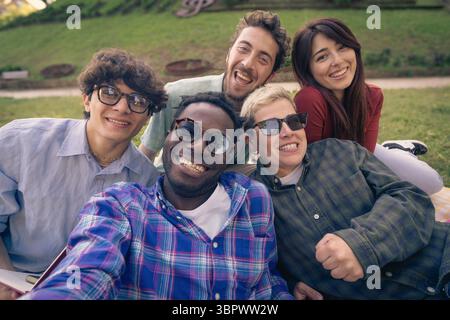 Un gruppo felice e variegato di giovani amici sorride e ride mentre si fa un energico selfie dal punto di vista all'aperto in un parco in una giornata di sole Foto Stock