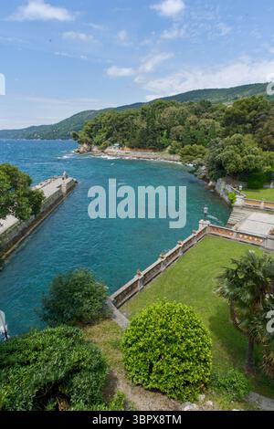 Una vivace insenatura blu adriatica con uno storico molo in pietra e lussureggianti giardini verdi, incorniciati da colline boscose sotto un luminoso cielo estivo. Foto Stock