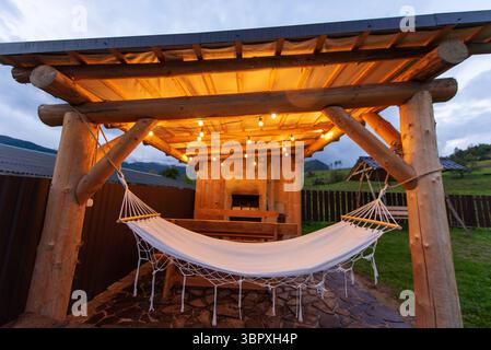 Un intimo pergolato esterno in legno con luci a corda e un'amaca bianca, adagiato in un'area erbosa con vista sulle montagne sullo sfondo. Foto Stock
