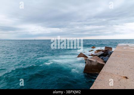 Un molo di cemento si estende in un mare instabile sotto un cielo nuvoloso, con onde che si infrangono contro grandi rocce. Foto Stock