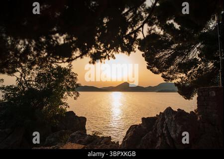 Vista del tramonto su un mare calmo incorniciato da alberi sagomati e costa rocciosa. Foto Stock