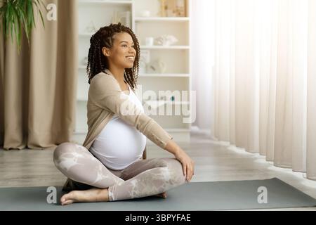 Donna incinta nera che esegue un tocco spinale sul tappetino da yoga, sorridendo con fiducia Foto Stock