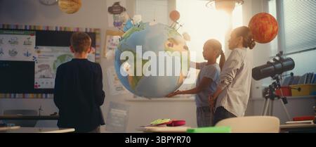 Lezione di scienze interattive: Insegnante femminile che educa i bambini intelligenti usando il modello della Terra. Scuola primaria i bambini che studiano energia rinnovabile e sistema solare in una moderna aula multietnica. Educazione STEM. Foto Stock