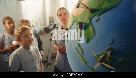 Classe Ecologia STEM: Scuola elementare ragazzo che attacca il modello in miniatura della turbina eolica a Globe. Insegnante donna che spiega la scienza e le fonti di energia rinnovabile agli studenti intelligenti in classe moderna. Foto Stock