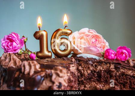 Dolce torta di compleanno al cioccolato di sedici anni decorata con 16 candele dorate illuminate e rose rosa. Torte fatta a mano per celebrare un evento speciale Foto Stock
