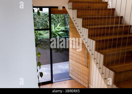 Scala galleggiante in legno con gradini aperti e ringhiera a cavo all'ingresso, rivelando il verde del patio. Moderno, minimalista, architettonico, interno, arredamento Foto Stock