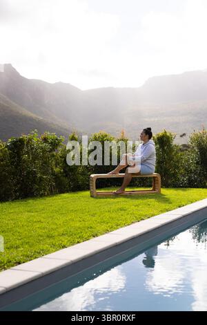 Donna asiatica di medie dimensioni seduta su una panchina di legno accanto alla piscina rettangolare su un prato lussureggiante, spazio fotocopie. Tranquillità, tempo libero, natura, paesaggio, benessere, all'esterno Foto Stock