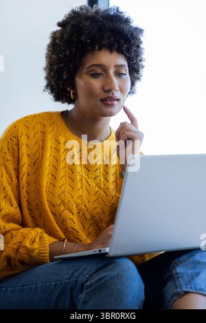 Donna che indossa un maglione giallo senape che lavora con cura su un notebook argentato a casa. Produttività, comfort, moderno, informale, spazio di lavoro, concentrati, tecnologia Foto Stock