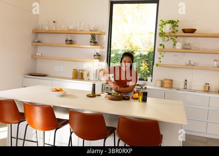 Donna asiatica in piedi a Kitchen Island condimento insalata in ciotola di legno con macinacaffè e olio d'oliva. Moderno, minimalista, culinario, stile di vita, interni, Foto Stock
