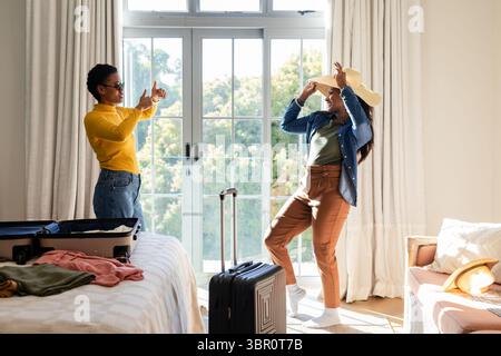 Diverse amiche che posano in camera da letto illuminata dal sole vicino a valigia aperta e cappello di paglia, spazio fotocopie. Stile di vita, amicizia, preparazione al viaggio, rilassato, vibr Foto Stock