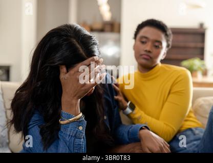 Diverse amiche che si siedono sul divano beige a casa tenendo la testa indossando braccialetti, offrendo la mano. Empatia, supporto, relax, accogliente, calore, amico Foto Stock