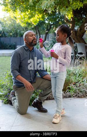 Padre e figlia afroamericani si inginocchiano nel patio del cortile posteriore soffiando bolle con bacchetta e tazza. Legami familiari, giochi all'aperto, tempo libero, natura, inno Foto Stock