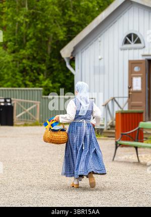 Sodertalje, Svezia - 6 giugno 2025: Una donna svedese in un tradizionale abito blu e bianco che porta un cesto intrecciato, che si allontana da un edificio in un ru Foto Stock