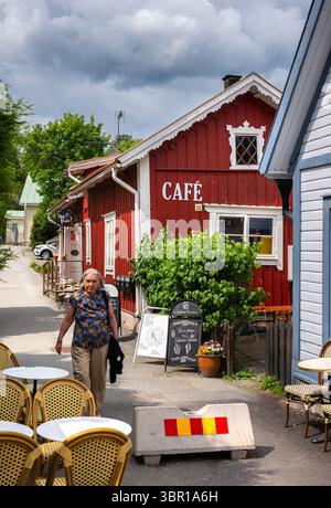 Sigtuna, Svezia - 9 giugno 2025: Un affascinante caffè con esterno in legno rosso e posti a sedere all'aperto. Una donna anziana passa accanto, circondata dalla lussureggiante greene Foto Stock