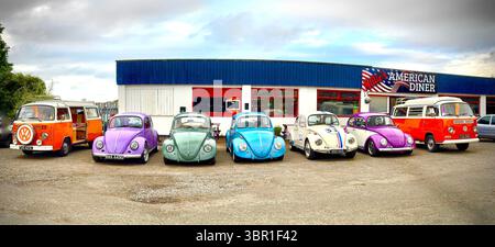 Panorama grandangolare della fila di Maggiolini Volkswagen d'epoca e furgoni camper vw T3 arancioni parcheggiati di fronte al cielo nuvoloso del ristorante americano Foto Stock