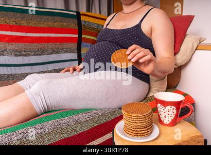 Donna incinta seduta sul divano di casa che si gusta biscotti, momento di maternità rilassato Foto Stock