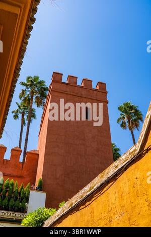 Torre di bastioni in terracotta e merlature incorniciate da mura ocra e palme ondeggianti sotto un cielo limpido di Siviglia Foto Stock