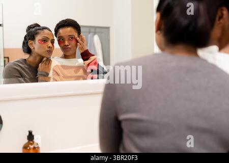 Madre indiana e figlia afro-americana che applicano cerotti sotto gli occhi allo specchio da trucco con bottiglia. Cura della pelle, bellezza, intimità, multigenerazionale, se Foto Stock