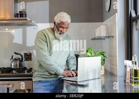Senior afroamericano che scrive su un computer portatile argentato nell'isola della cucina vicino a smartphone, impianto in vaso. Professionale, moderno, minimalista, stile di vita, ho Foto Stock