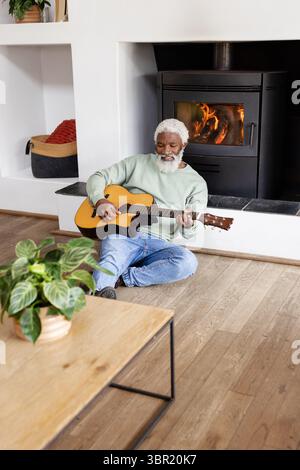 Uomo afroamericano senior che suona la chitarra nel soggiorno con tavolino da caffè, pianta e fornello. Musicista, relax, accogliente, campagna, acustica, no Foto Stock