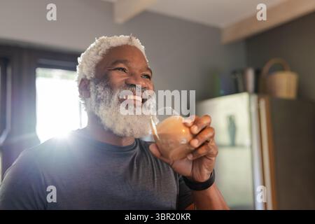 Senior uomo afroamericano sorridente indossando uno smartwatch, tenendo la tazza di caffè schiumosa in una cucina illuminata dal sole. Anziani, benessere, ospitalità, stile di vita moderno Foto Stock